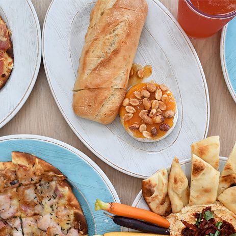 A feast of pizzas, a baguette with almond cookies, hummus, pita crisps, wine, and a citrus drink on blue and white plates.