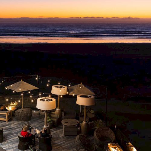 People dine on a dusk-lit beach deck with umbrellas, string lights, and warm fire pits, overlooking a calm ocean at sunset.