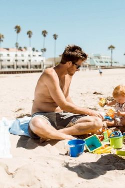 A family builds sandcastles on the beach: dad, mom, and a child play with colorful buckets and toys near the shoreline and a sunny promenade.