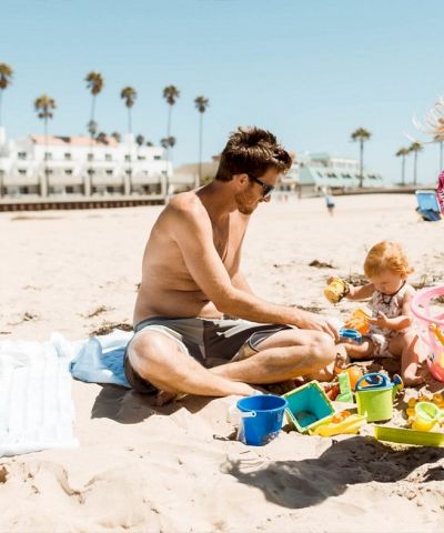 A family builds sandcastles on the beach: dad, mom, and a child play with colorful buckets and toys near the shoreline and a sunny promenade.