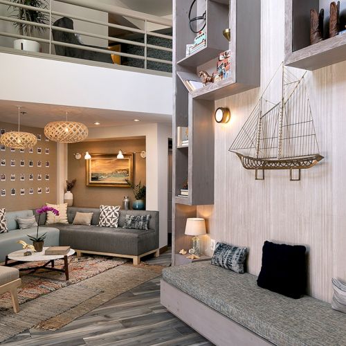 A cozy, stylish living room with a gray sectional, matching cushions, and a bay window nook; nautical decor, warm lighting, and contemporary shelves.