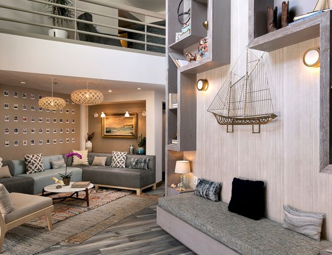 A cozy, stylish living room with a gray sectional, matching cushions, and a bay window nook; nautical decor, warm lighting, and contemporary shelves.