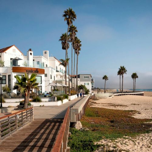 Coastal beach town with white buildings, palm trees, a boardwalk along the shore, and a sandy beach meeting the calm blue sea.