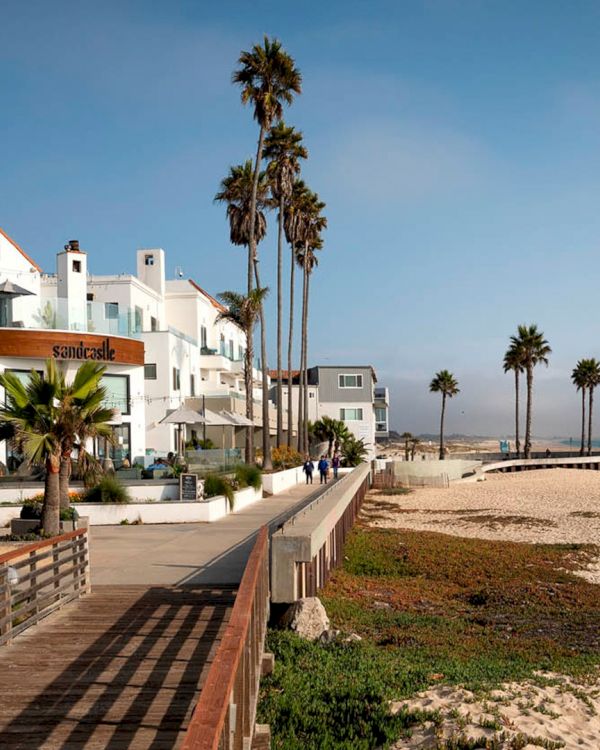 Coastal beach town with white buildings, palm trees, a boardwalk along the shore, and a sandy beach meeting the calm blue sea.