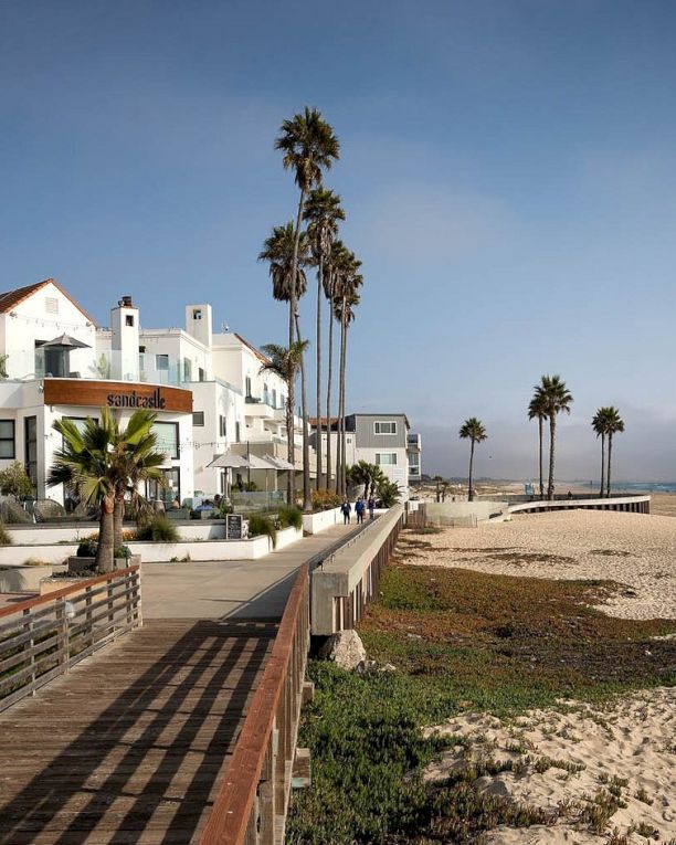 A sunny coastal scene with white buildings, palm trees, a wooden boardwalk, and a sandy beach meeting the calm ocean.
