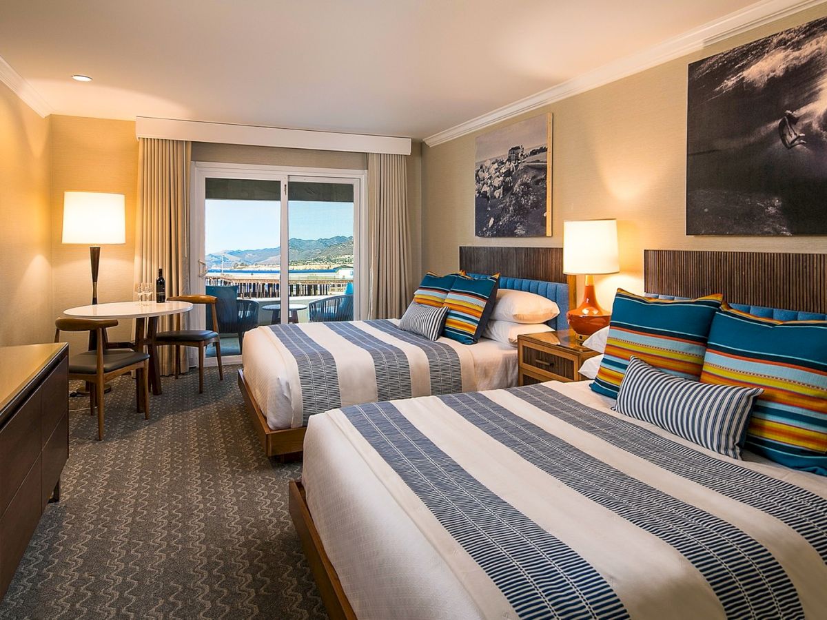 Two twin beds with striped blue-gray linens and bright, colorful pillows in a modern hotel room, balcony view, warm lighting, and large wall art.