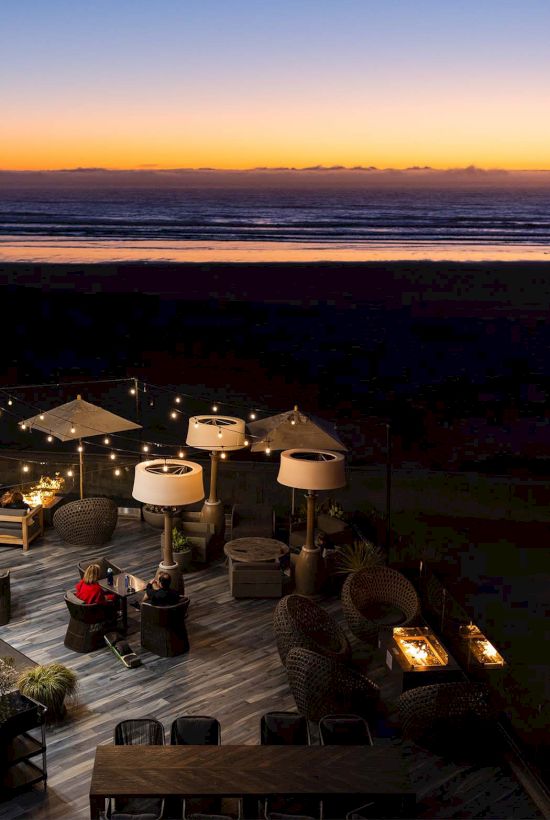 Beachfront sunset scene with a lit outdoor seating deck, umbrellas, string lights, and a calm ocean glow as dusk settles.