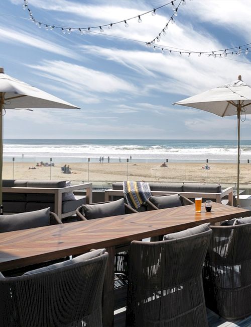 Beachfront dining deck with wicker tables, umbrellas, string lights, and a view of the ocean and sandy shore under a blue sky. Ending this sentence.