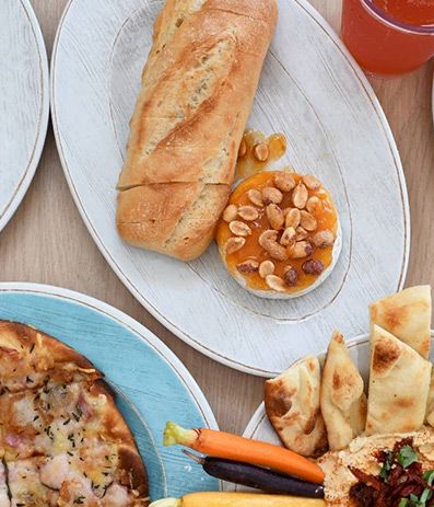 A spread of pizzas on colorful plates, plus a baguette with almonds, naan-style chips, drinks, and dipping sauces on a wooden table.