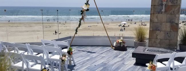 A small beach wedding setup with a triangular arch decorated with flowers, white chairs facing the ocean, and a ceremony area on a wooden deck by the shore.