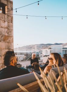 A couple sits on a bench in a sunny courtyard with potted plants, warm stone walls, and a tower in the background.