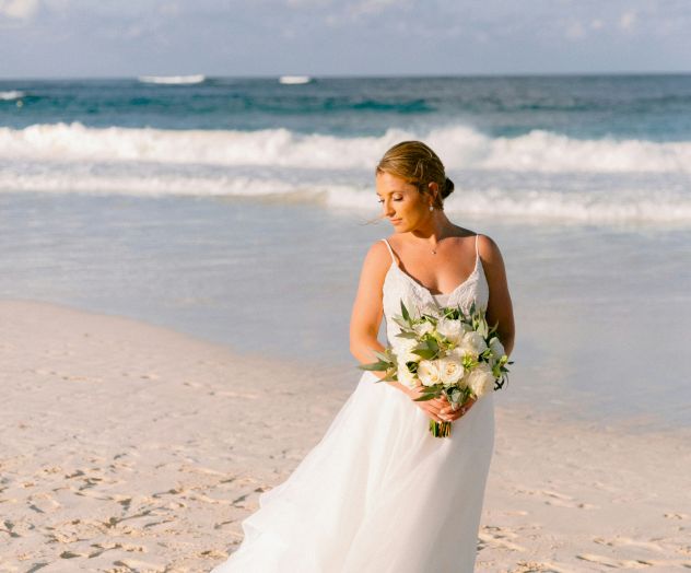 A bride in a white gown stands on a sandy beach, holding a bouquet, with the ocean and waves in the background.