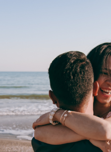 Two people hug on a sunny beach, smiling at the camera, with the ocean and waves in the background.