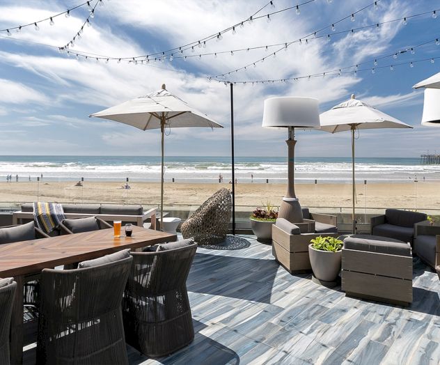 A sunny outdoor cafe on a wooden deck by the water with tables, chairs, and sailboats in the distance, under a blue sky with clouds.