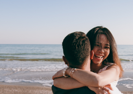 Two people hug warmly on a sandy beach with the ocean and a clear sky in the background, sharing a joyful moment together.