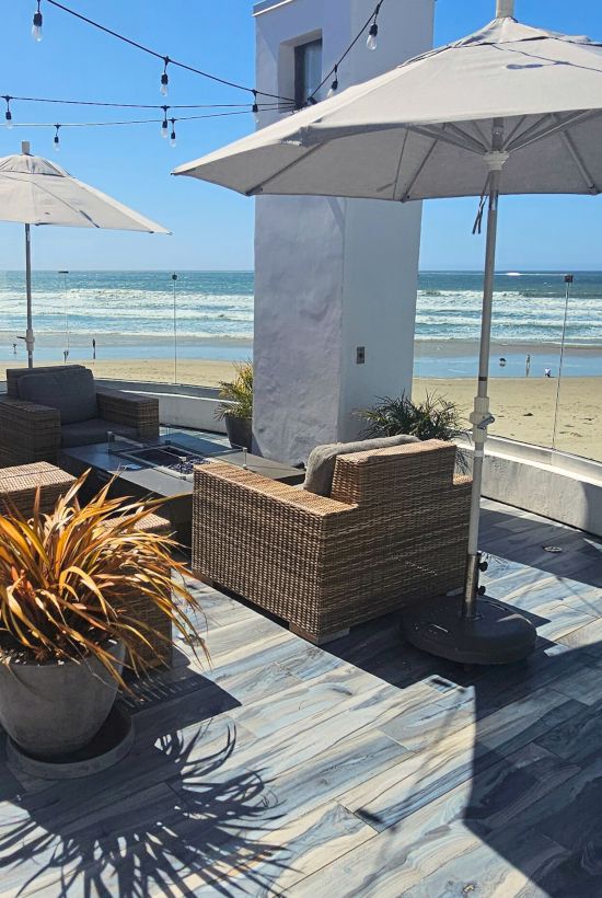 A sunny beachside deck with wicker furniture, umbrellas, glass railing, potted plants, and views of the ocean and surfers beyond.