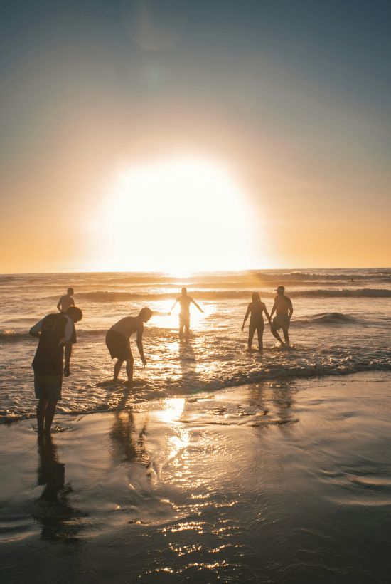 People playing at the shore during a bright sunset, silhouettes dancing in shallow water along a reflective beach.